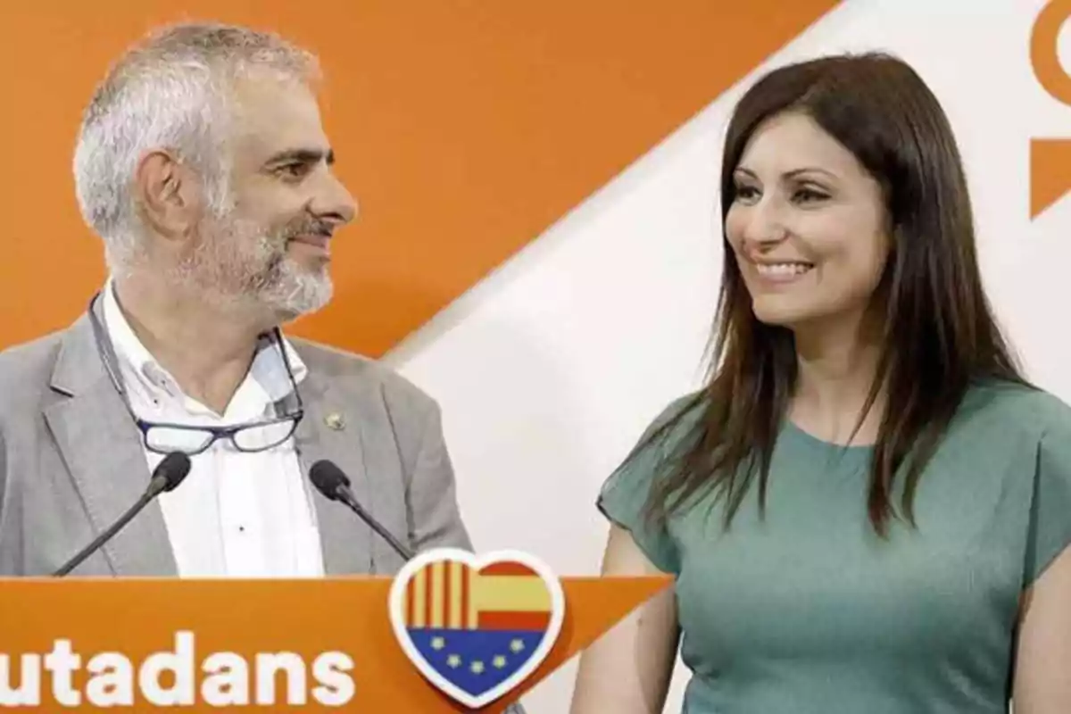 Dos personas sonrientes en un evento con un fondo naranja y blanco y un cartel que dice "utudans" con un corazón que contiene una bandera.