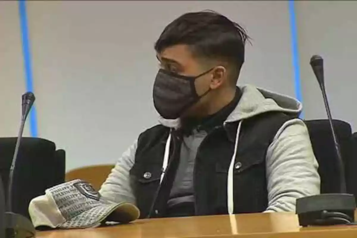 Un hombre con mascarilla negra y chaqueta de mezclilla con capucha gris está sentado en una sala de conferencias o tribunal, con un micrófono frente a él y una gorra sobre la mesa.