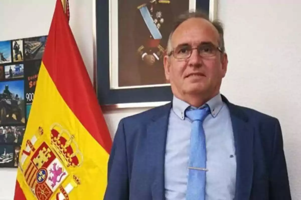 Hombre con traje y corbata azul posando junto a una bandera de España y un cuadro en la pared.