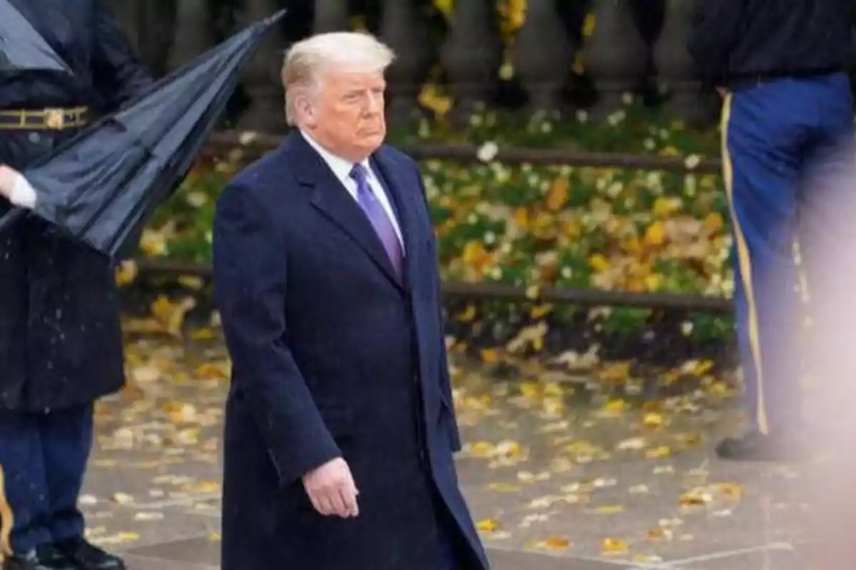 Un hombre con abrigo oscuro y corbata morada camina al aire libre en un entorno otoñal, mientras una persona detrás de él sostiene un paraguas.