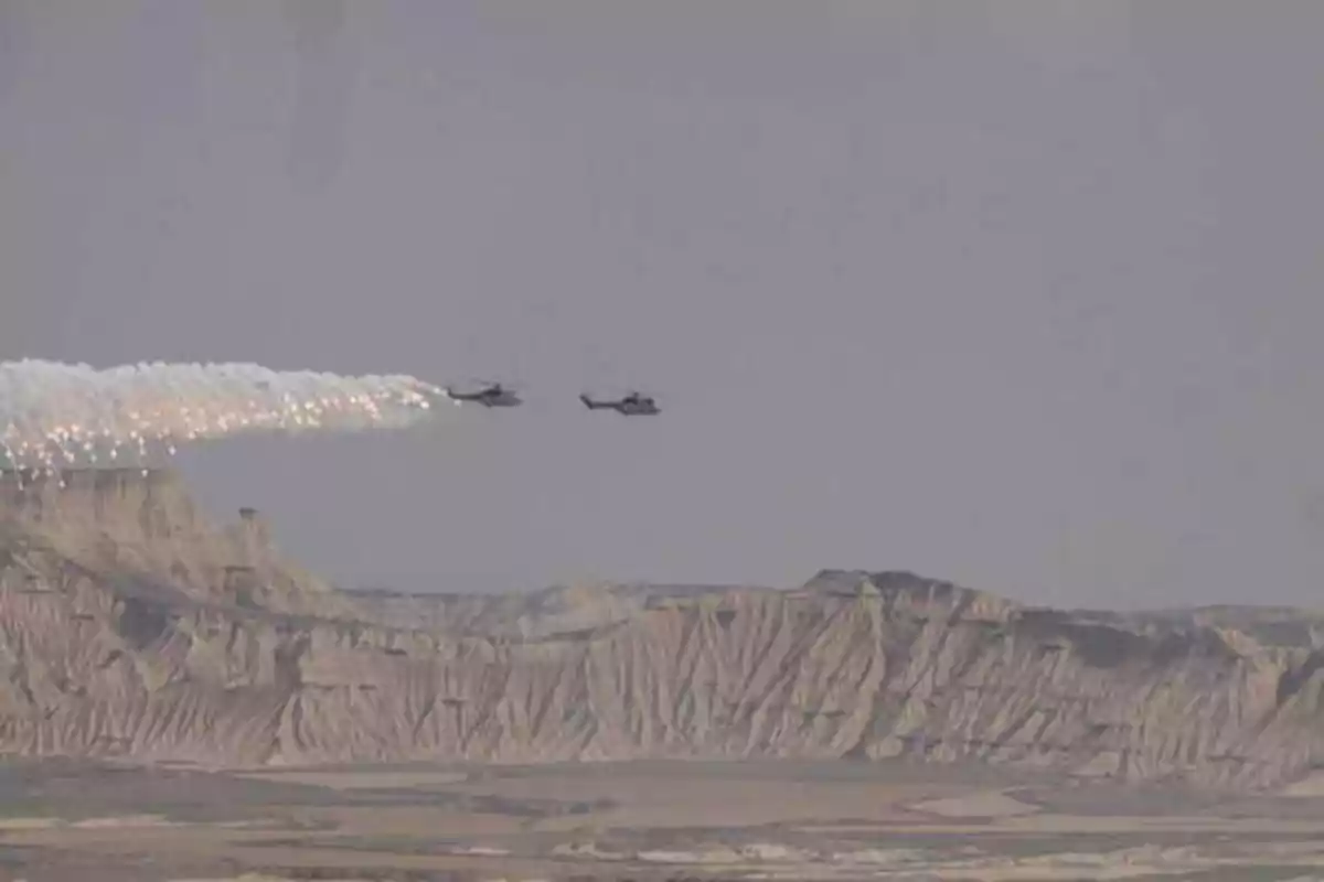 Dos helicópteros militares volando sobre un paisaje montañoso árido con uno de ellos lanzando bengalas.