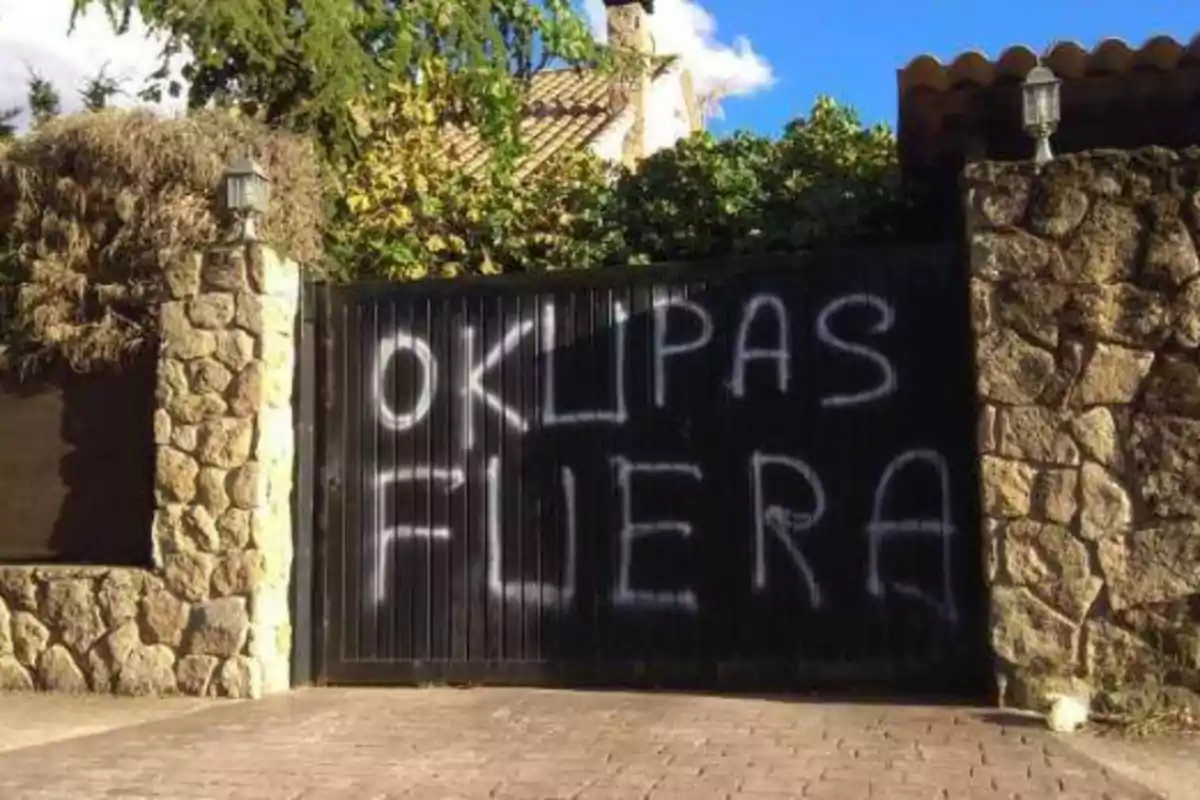 Una puerta de garaje negra con grafiti blanco que dice "OKUPAS FUERA", flanqueada por dos columnas de piedra y rodeada de vegetación.