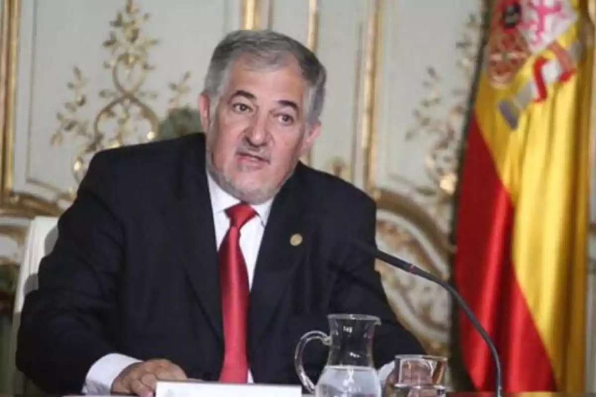 Hombre de traje oscuro y corbata roja hablando en una conferencia con una bandera de España en el fondo.