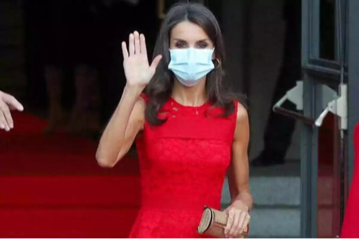 Persona con vestido rojo y mascarilla saludando con la mano.