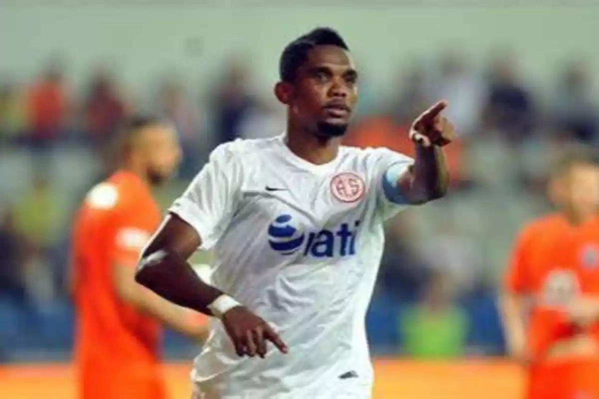 Un jugador de fútbol con uniforme blanco señala con el dedo mientras está en el campo de juego, con otros jugadores y espectadores desenfocados en el fondo.