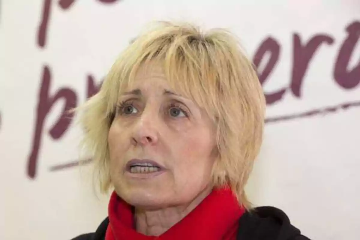 Una mujer de cabello rubio corto y bufanda roja hablando frente a un fondo con letras borrosas.