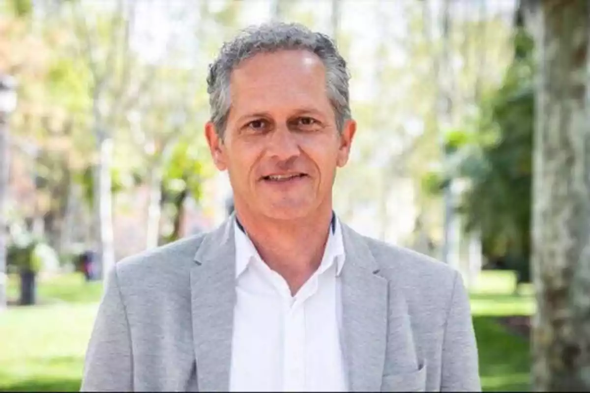 Hombre de cabello canoso con chaqueta gris y camisa blanca sonriendo en un parque.