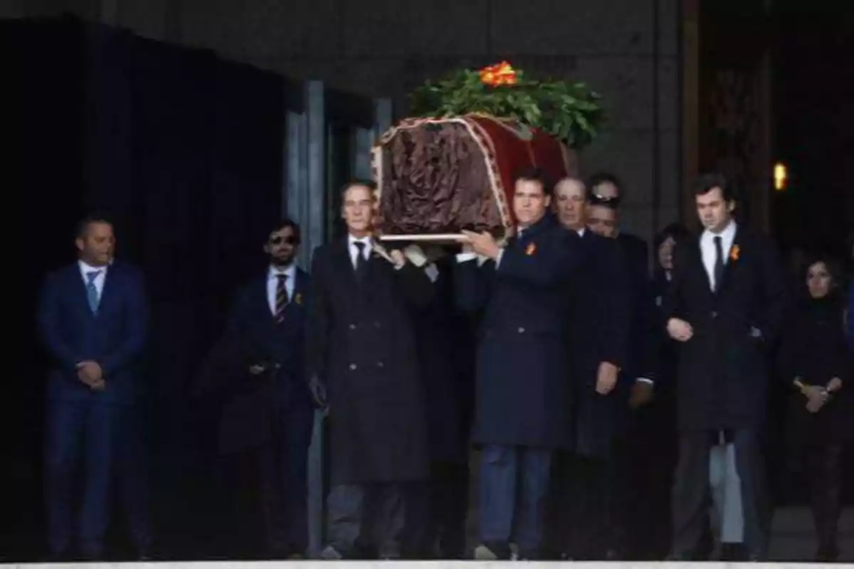 Un grupo de personas vestidas de traje y abrigo oscuro lleva un ataúd cubierto con una tela roja y decorado con flores.