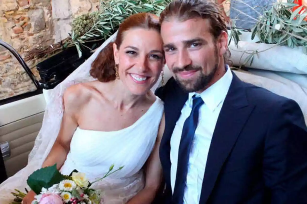 Una pareja sonriente vestida de novios, la mujer con un vestido blanco y el hombre con un traje oscuro, posando juntos en un entorno rústico.