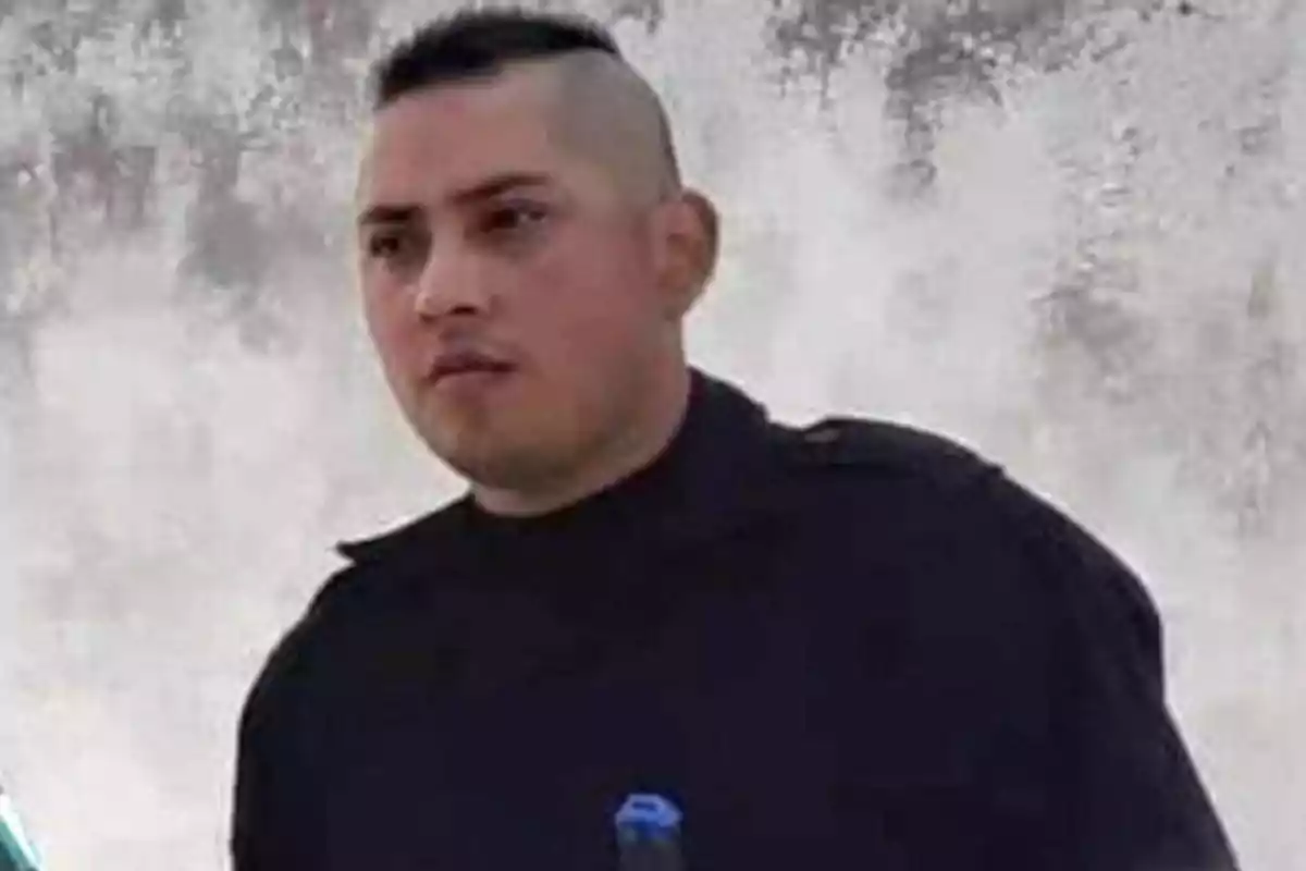 Hombre con uniforme negro y corte de cabello militar frente a una pared gris.