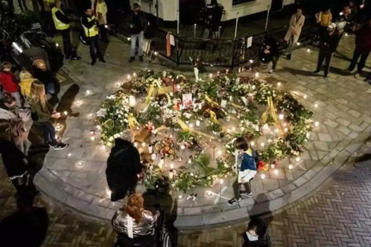 Personas reunidas alrededor de un memorial circular con velas y flores en una calle pavimentada.