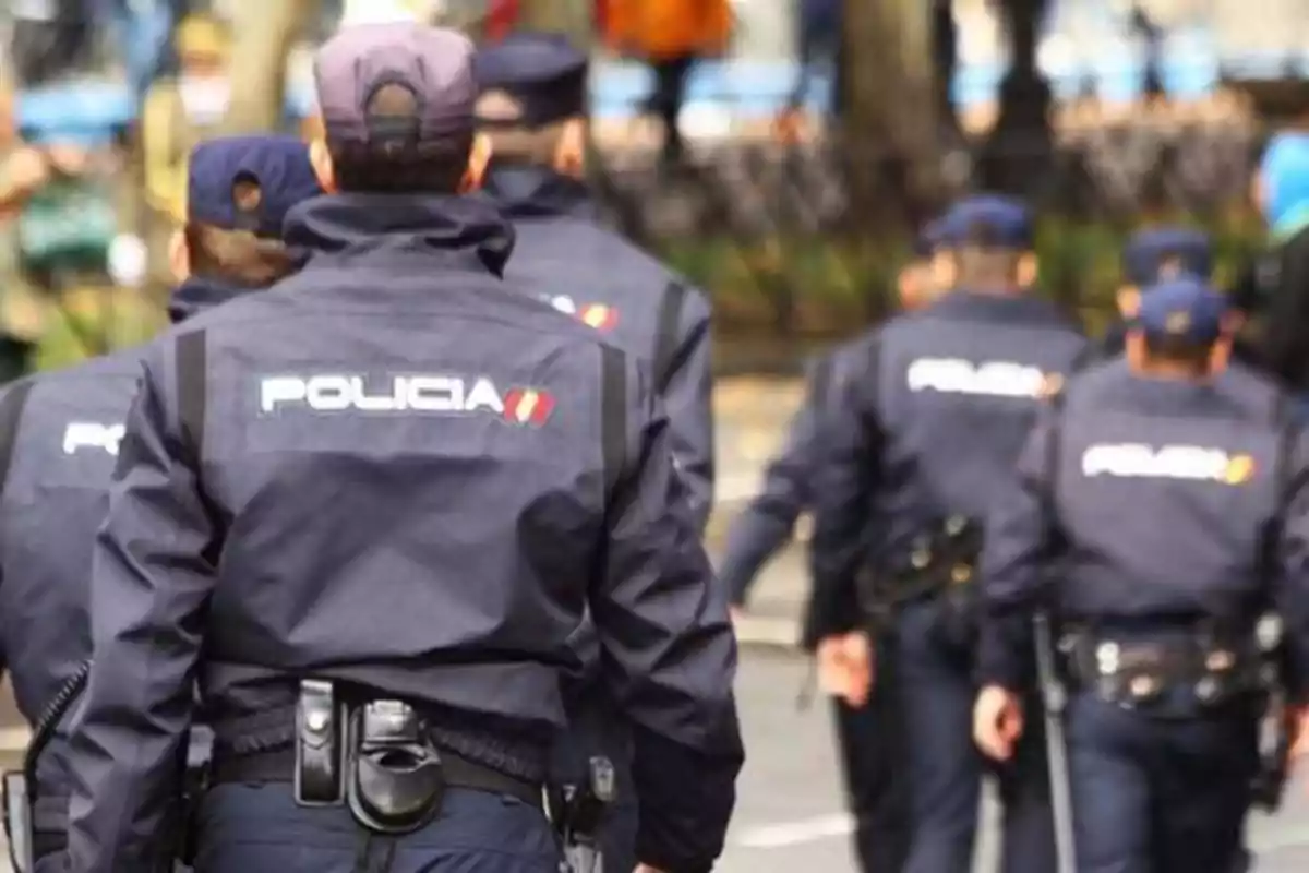 Un grupo de policías caminando en fila, vistos desde atrás, con uniformes oscuros y la palabra "POLICÍA" visible en sus chaquetas.