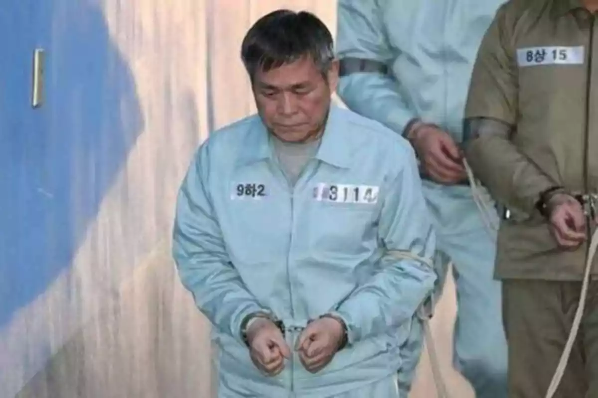 Un hombre con uniforme de prisionero y esposado, caminando junto a otros prisioneros.