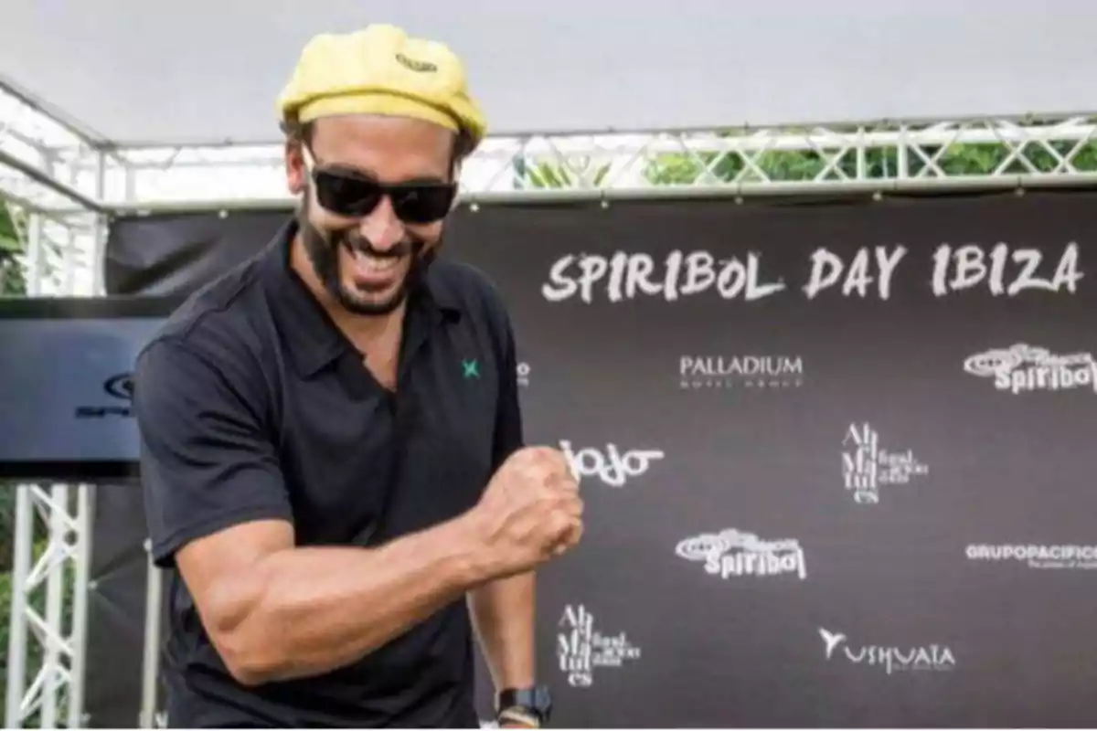 Un hombre con gafas de sol y una boina amarilla sonríe y hace un gesto de victoria con el puño en un evento llamado "Spiribol Day Ibiza".