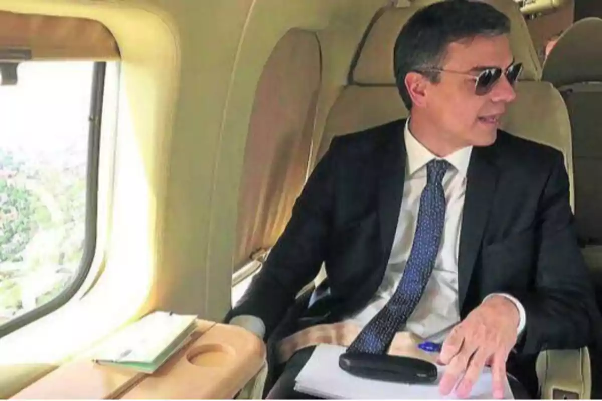 Hombre con traje y corbata, usando gafas de sol, sentado en un avión privado, con una carpeta y un bolígrafo sobre la mesa.