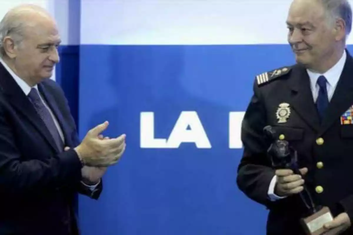 Dos hombres, uno de ellos en uniforme militar, están en un evento; uno de ellos sostiene un trofeo mientras el otro aplaude.