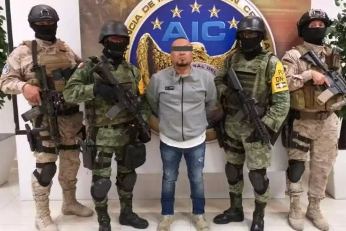 Cinco hombres, cuatro de ellos con uniformes militares y armas, posan junto a un hombre con los ojos cubiertos frente a un emblema de la Agencia de Investigación Criminal (AIC).
