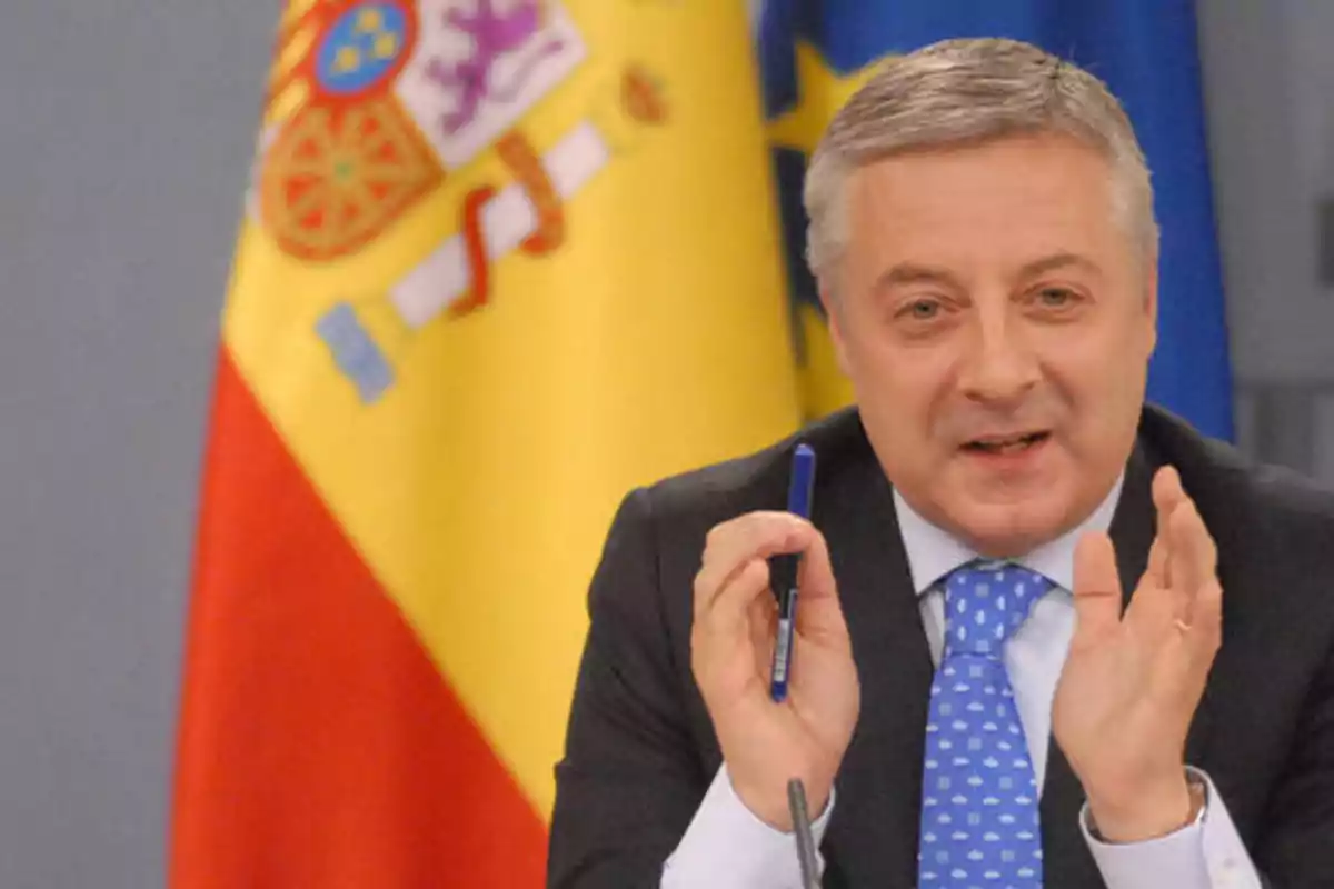 Hombre de traje y corbata azul con puntos blancos, sosteniendo un bolígrafo, hablando frente a un micrófono con la bandera de España de fondo.