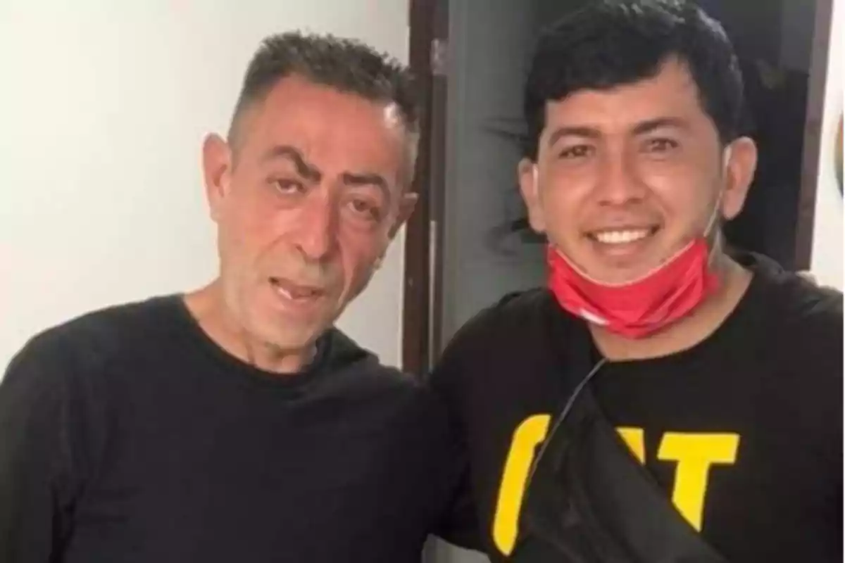 Dos hombres posando para una foto, uno con camiseta negra y el otro con camiseta negra y mascarilla roja en el cuello.