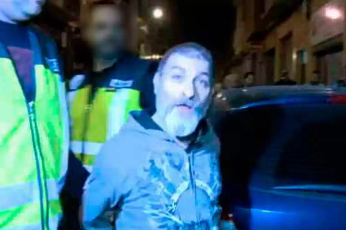 Un hombre con barba y cabello corto es escoltado por dos personas con chalecos reflectantes en una calle durante la noche.