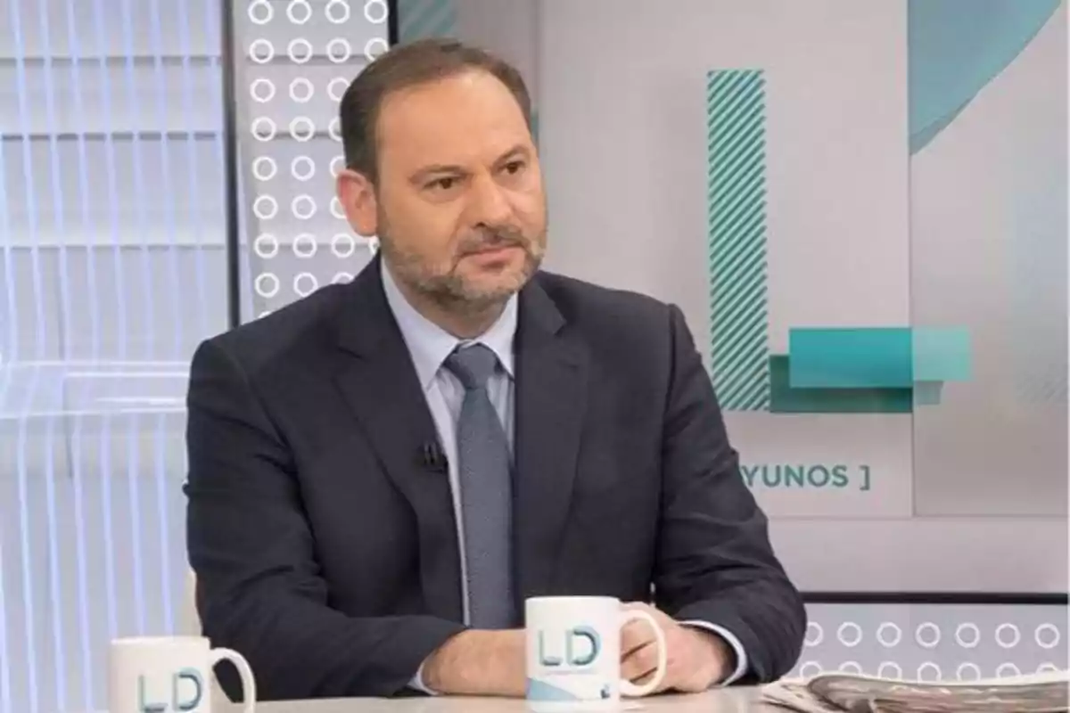 Un hombre con traje y corbata está sentado en un estudio de televisión sosteniendo una taza blanca con las letras LD.