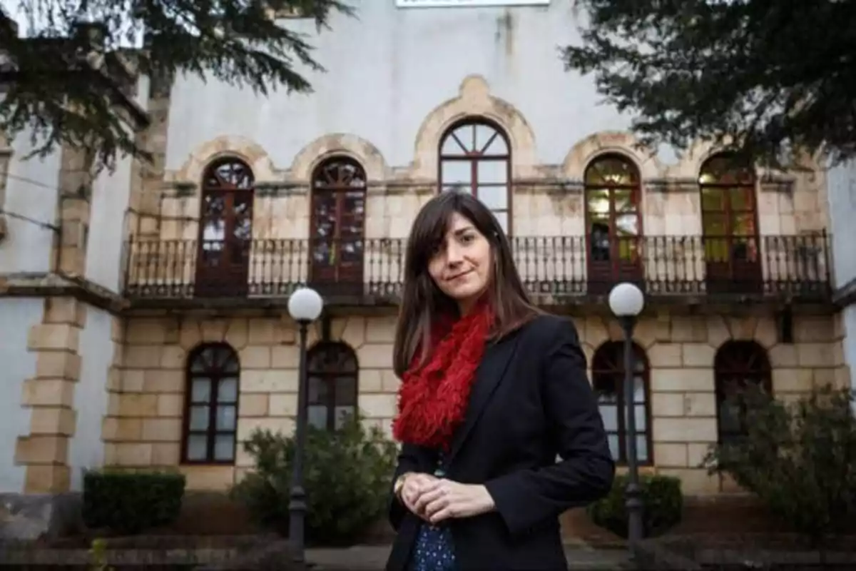 Una mujer con bufanda roja y chaqueta negra está de pie frente a un edificio antiguo con ventanas arqueadas y balcones de hierro forjado.