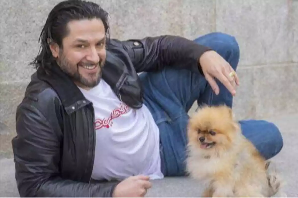 Un hombre sonriente con chaqueta de cuero y camiseta blanca está recostado en el suelo junto a un pequeño perro de raza Pomerania.