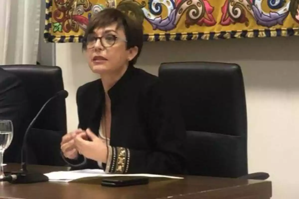 Una mujer con gafas y chaqueta negra está sentada en una mesa, hablando frente a un micrófono, con un fondo decorativo colorido detrás de ella.