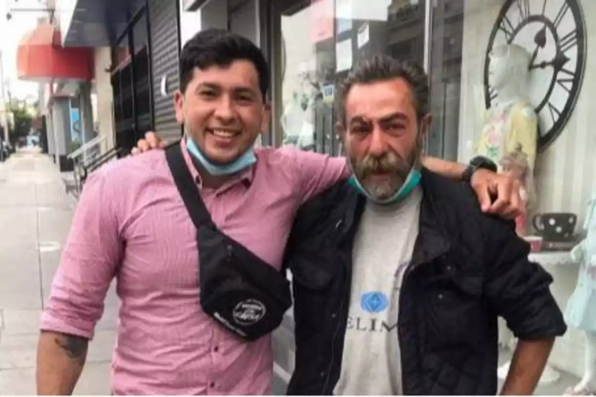 Dos hombres posando juntos en una acera, ambos con mascarillas bajadas y sonriendo.