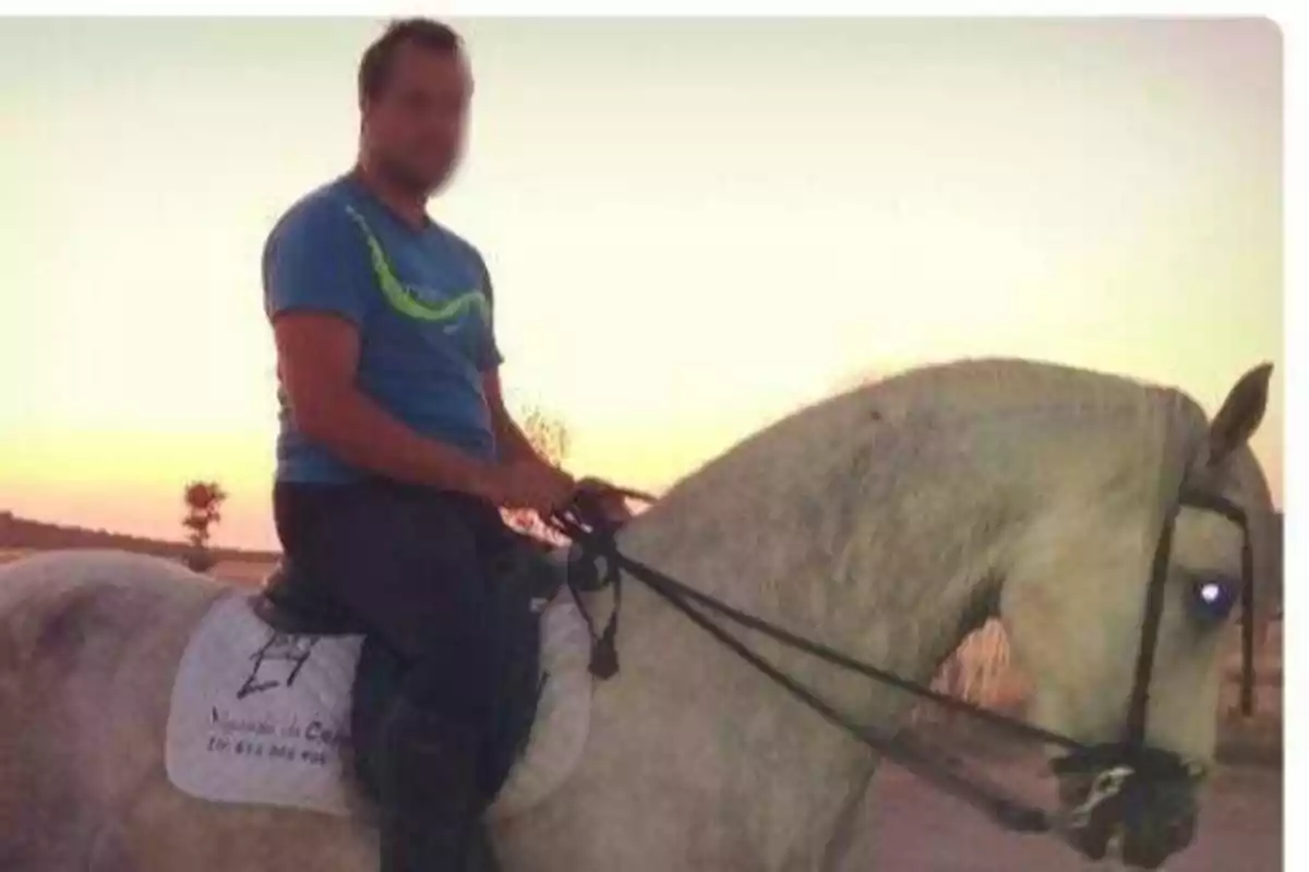 Un hombre con una camiseta azul está montando un caballo blanco al atardecer.