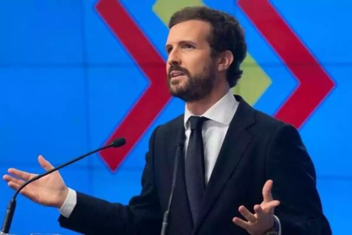 Hombre con traje y corbata hablando en un podio con micrófonos frente a un fondo azul con flechas rojas y amarillas.