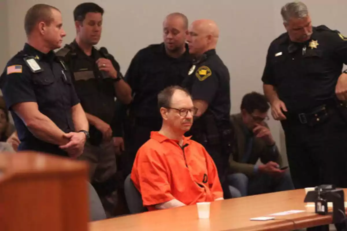 Un hombre con uniforme naranja está sentado en una sala de tribunal rodeado de varios oficiales de policía.