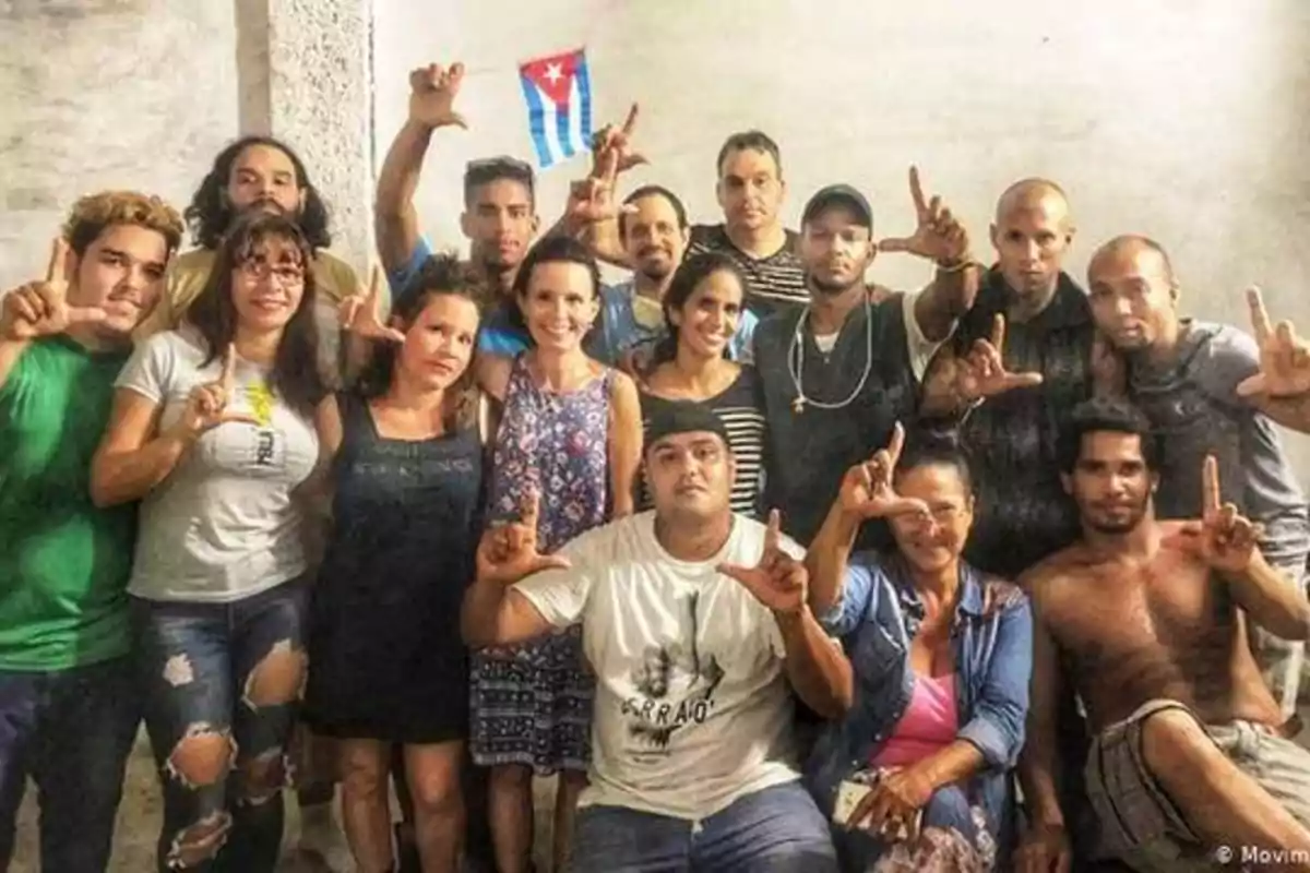 Un grupo de personas posando para una foto, algunas de ellas haciendo el gesto de la letra "L" con sus manos, con una bandera cubana en el fondo.