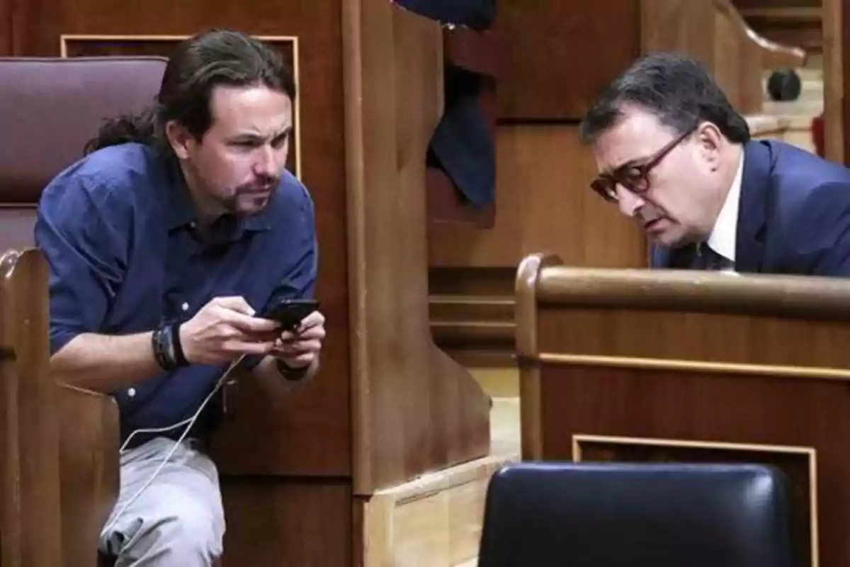 Dos hombres conversando en un entorno de oficina, uno de ellos revisando su teléfono móvil.