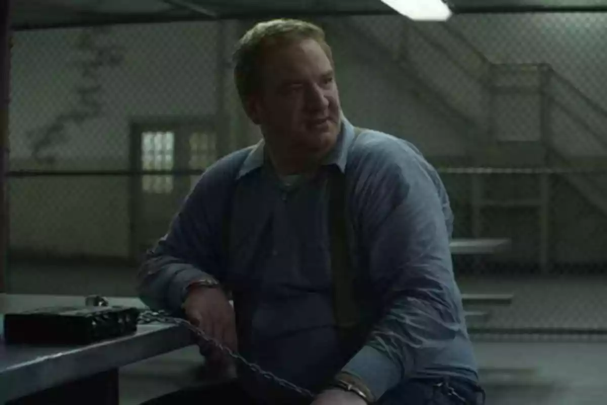 Un hombre con camisa azul está esposado y encadenado a una mesa en lo que parece ser una sala de interrogatorios o una prisión.