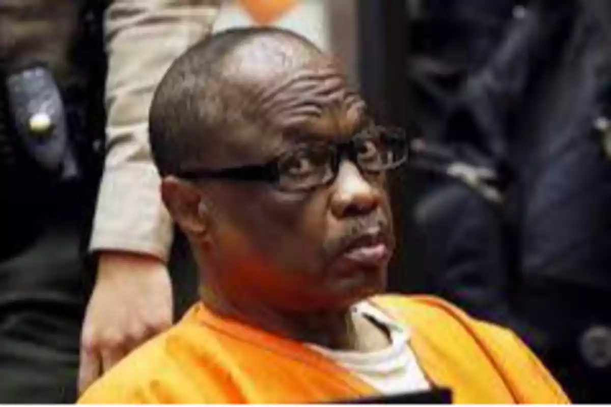 Un hombre con gafas y uniforme naranja sentado en una sala de tribunal.