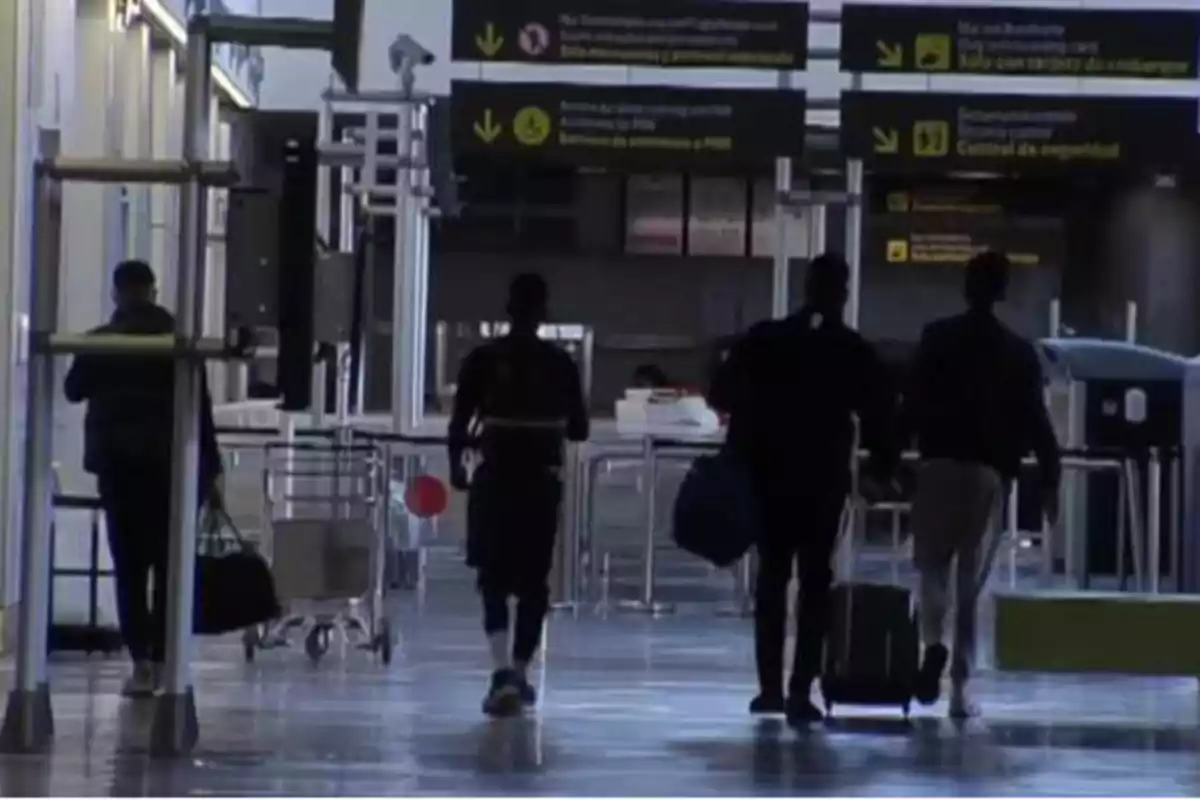 Personas caminando en un aeropuerto con maletas y señales de dirección en el fondo.