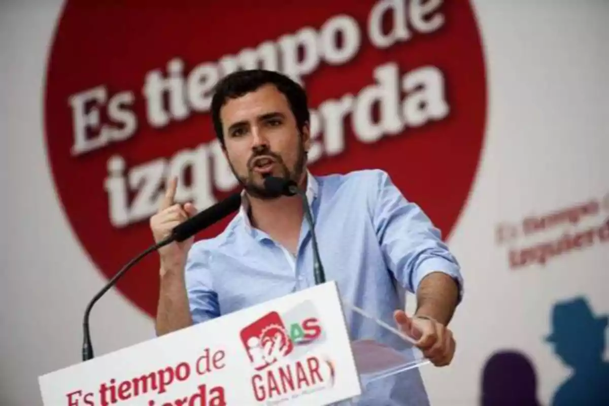 Un hombre con barba y camisa azul está dando un discurso en un podio con un micrófono, detrás de él hay un cartel rojo con el texto "Es tiempo de izquierda".