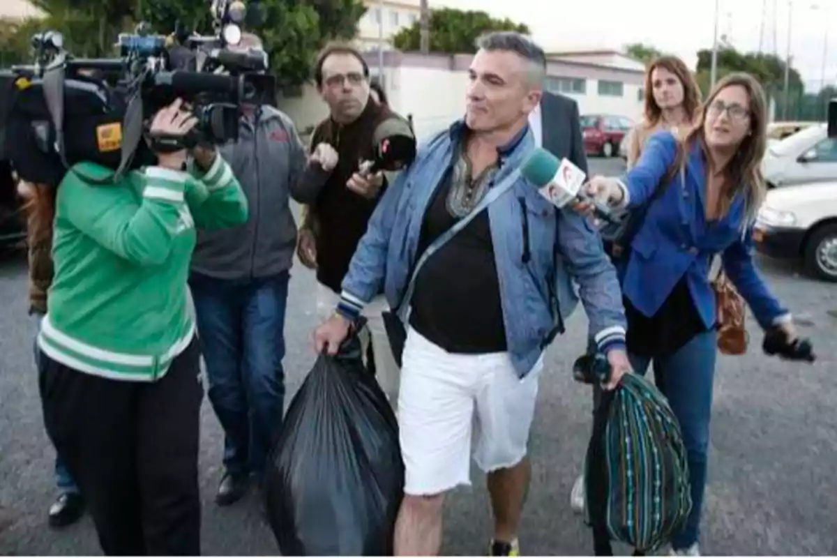 Un hombre camina por la calle llevando dos bolsas mientras es rodeado por periodistas y camarógrafos que lo graban y le hacen preguntas.