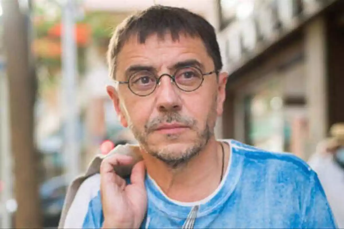 Hombre con gafas redondas y barba corta, vestido con una camiseta azul, mirando a la cámara en un entorno urbano.