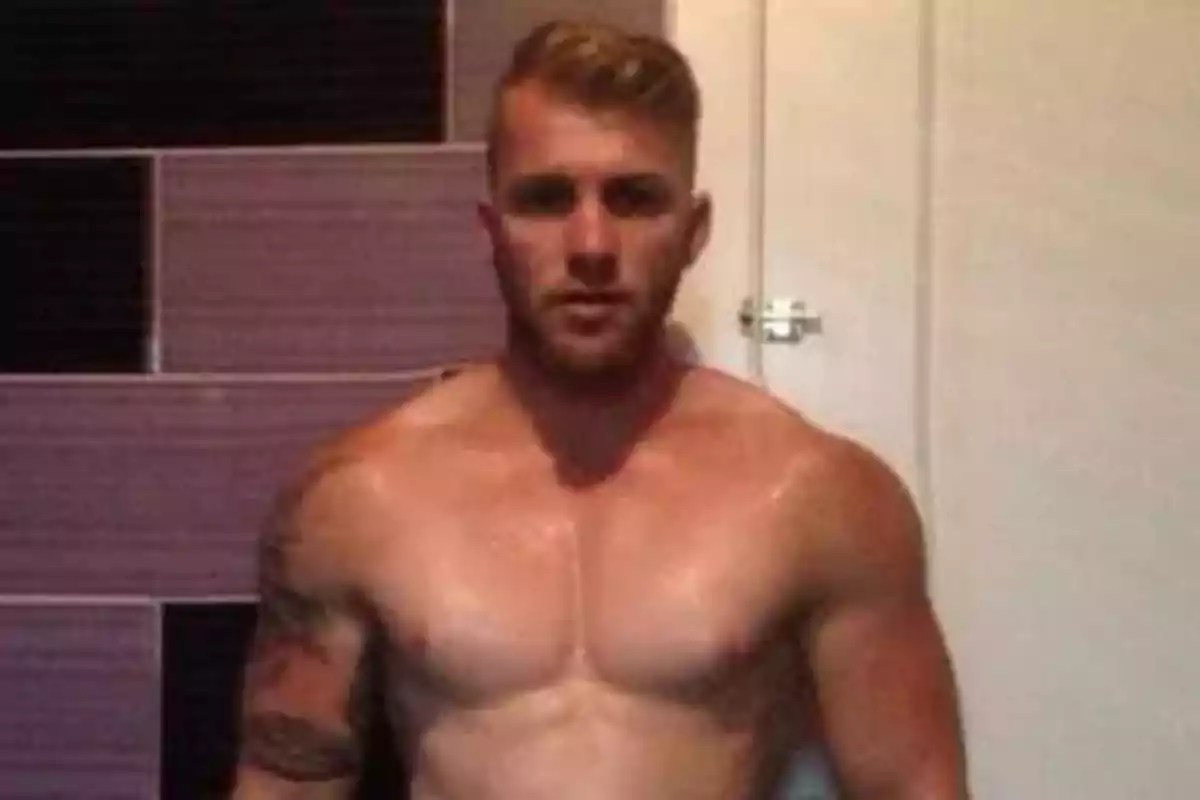 Hombre musculoso sin camisa con tatuajes en el brazo izquierdo, de pie frente a una pared con azulejos morados y negros.
