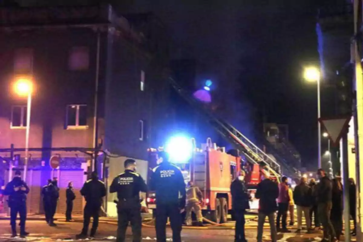 Policías y bomberos trabajando en la escena de un incendio en un edificio durante la noche, con luces de emergencia iluminando el área y varias personas observando.
