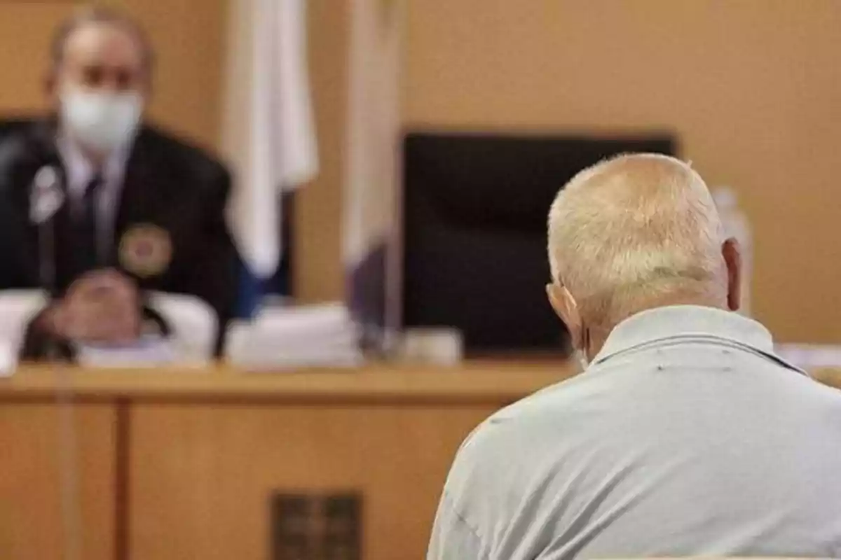 Un hombre mayor de espaldas en una sala de tribunal frente a un juez con mascarilla.