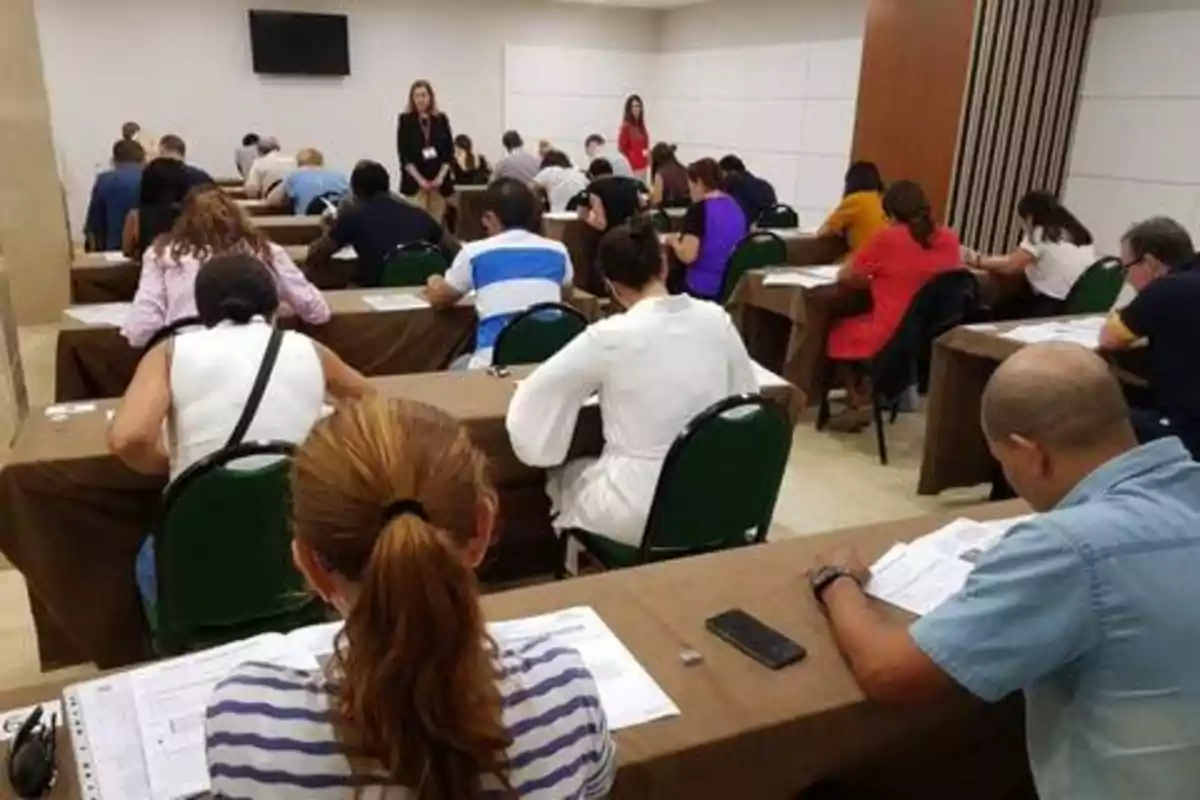 Personas sentadas en un salón de clases tomando un examen mientras dos personas de pie supervisan.