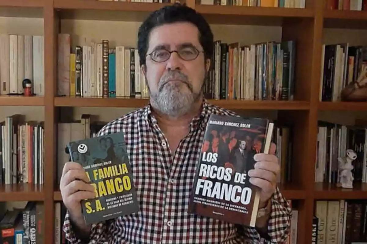 Un hombre con gafas y barba sostiene dos libros, uno en cada mano, frente a una estantería llena de libros.
