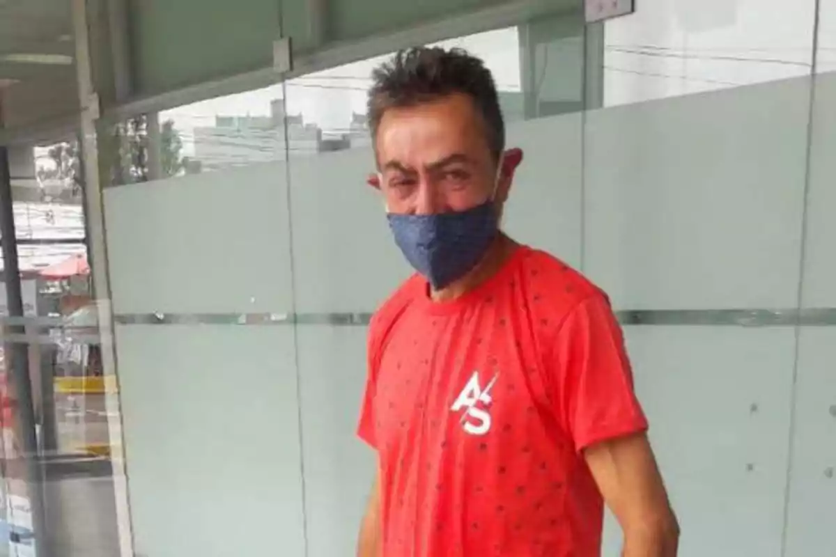 Hombre con camiseta roja y mascarilla azul de pie frente a una pared de vidrio.