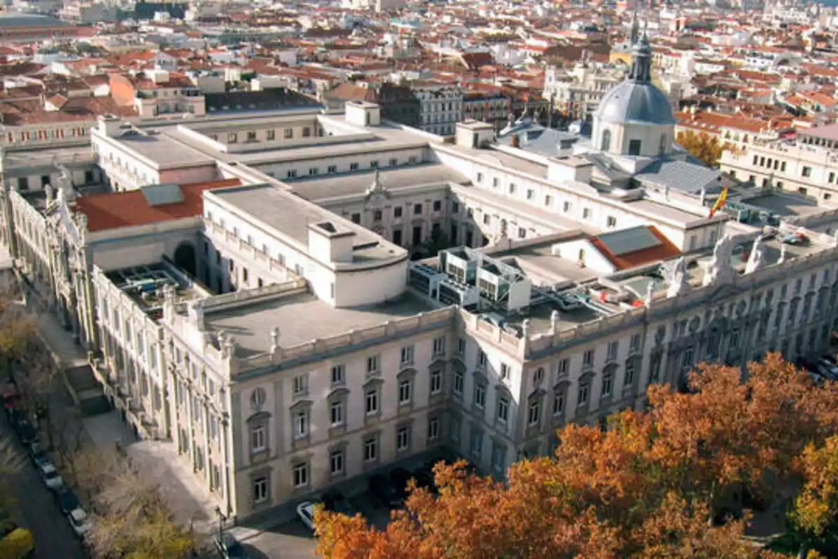 Vista aérea de un gran edificio histórico con una cúpula y múltiples alas, rodeado de una ciudad con techos de tejas rojas y árboles en primer plano.