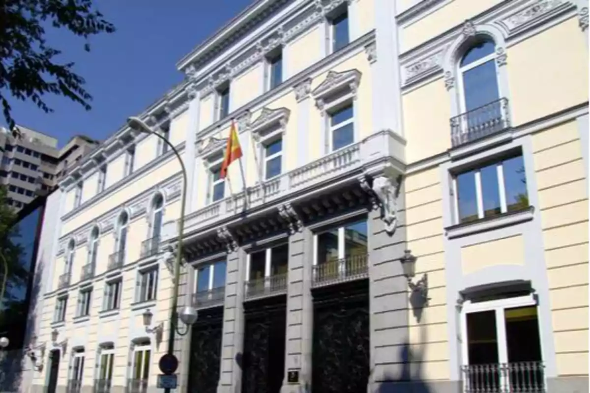 Edificio histórico con fachada blanca y detalles arquitectónicos clásicos, con una bandera de España ondeando en el centro.