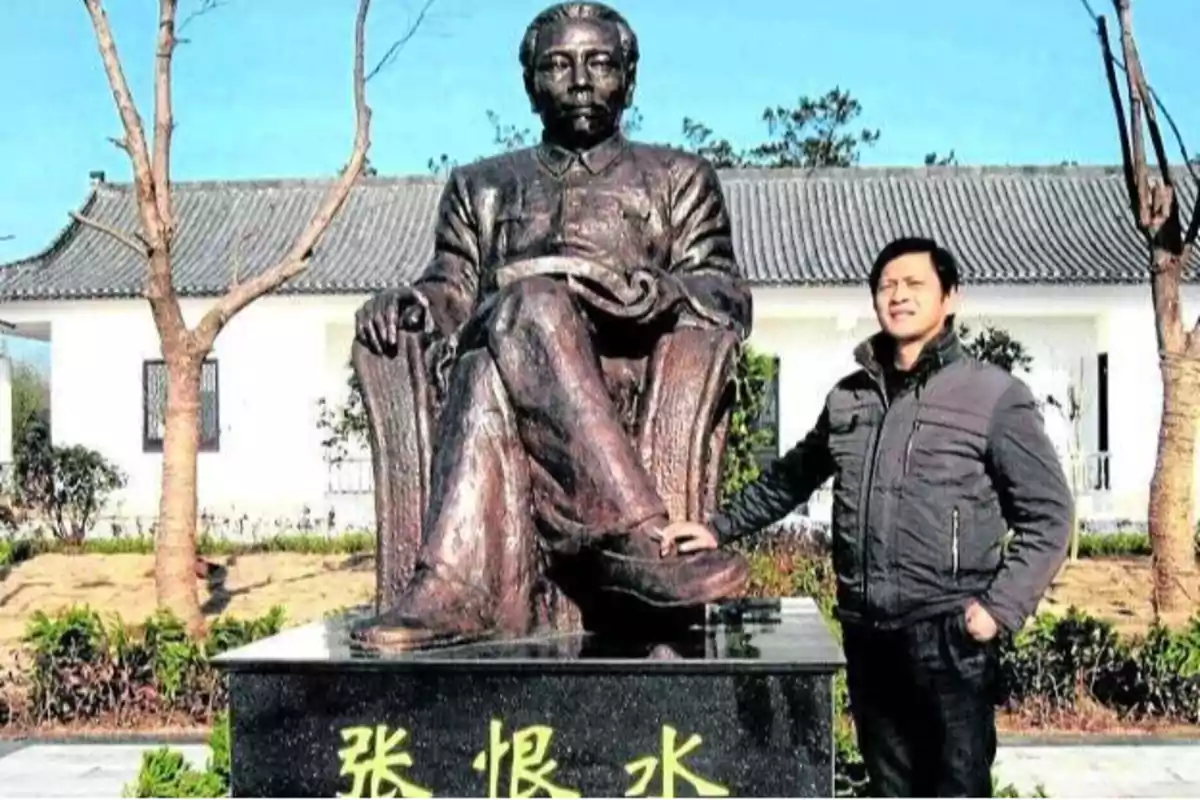 Un hombre posando junto a una estatua de bronce en un jardín con árboles y una casa de fondo.