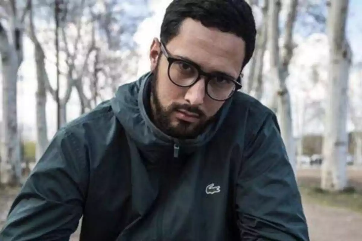 Hombre con gafas y barba, vistiendo una chaqueta oscura, en un entorno al aire libre con árboles de fondo.